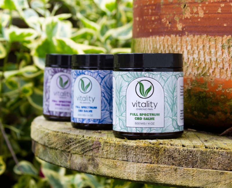 Three containers of Vitality's CBD salves for chronic pain, relaxation, and tension on a wooden shelf with some leaves in the background. 