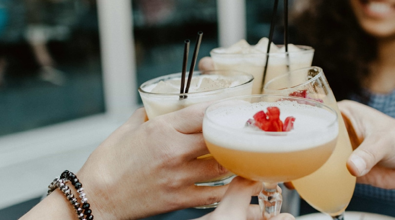 Four hands, each holding a cocktail and toasting with each other.